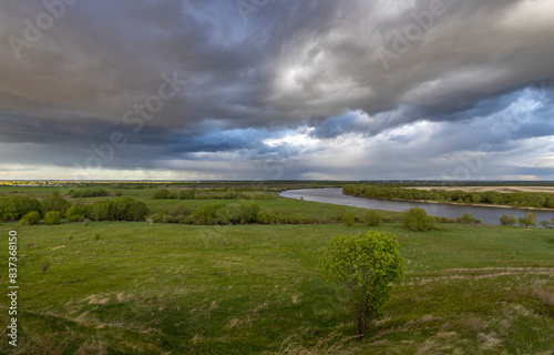 rural landscape with a meandering river cutting through a vast green meadow. The horizon is dominated by a dramatic sky filled with storm clouds  suggesting an impending change in weather.