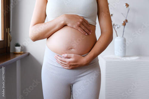 Pregnant Unrecognized Woman Dressed In A Sporty Outfit Holding Her Belly . Expecting Child. Home Indoor Interior. Concept Of Pregnancy and New Life.