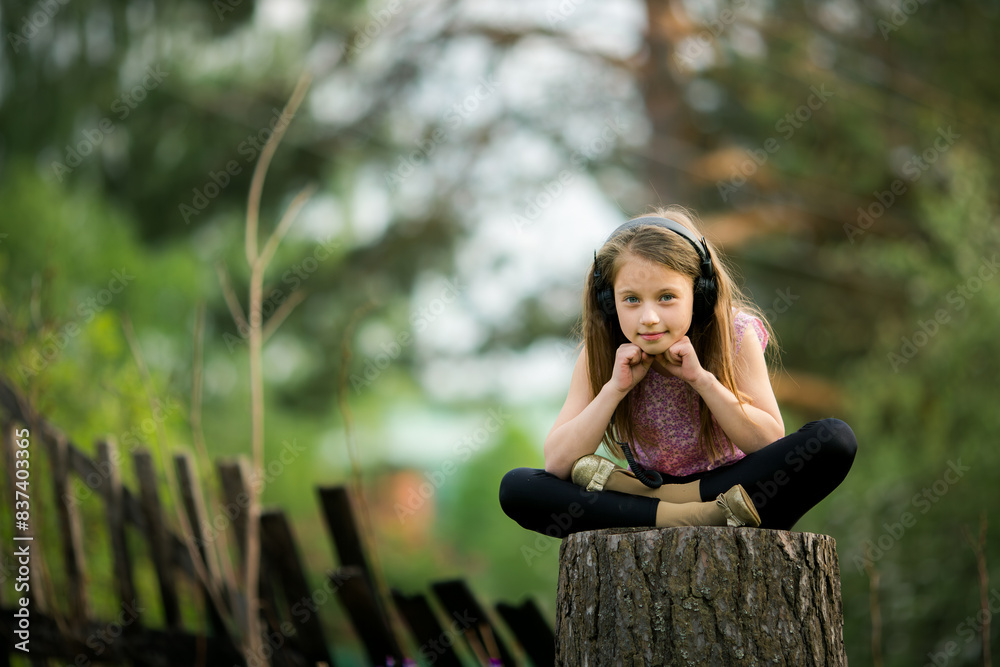 A teenage girl in headphones sits on a tree stump in a village, enjoying a moment of solitude. A teenage girl in headphones sits on a tree stump in a village, enjoying a moment of solitude.