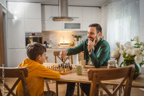 Father with cup of hot drink and son play chess together at home