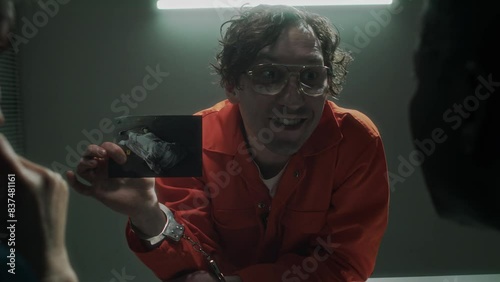 Medium shot of detectives interrogating psychotic murder suspect in orange jumpsuit, who is taunting them with maniac laughter, shaking photo of victim body, shouting, then lighting cigarette