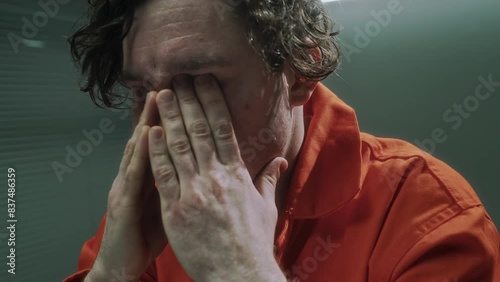 Close-up shot of young Caucasian male serial murder suspect in orange prison jumpsuit sitting in interrogation room in pretrial detention facility, rubbing eyes while considering crime confession photo