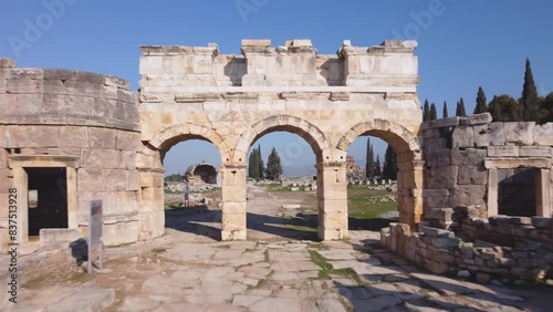 Wallpaper Mural Historic arched city gates to the ruins on Main Street promenade walkway. Fly UP to Wide Landscape view Ancient Roman, Greek and now Turkish city of Hierapolis. Pamukkale, Denizli, Turkey (Türkiye) 4K Torontodigital.ca
