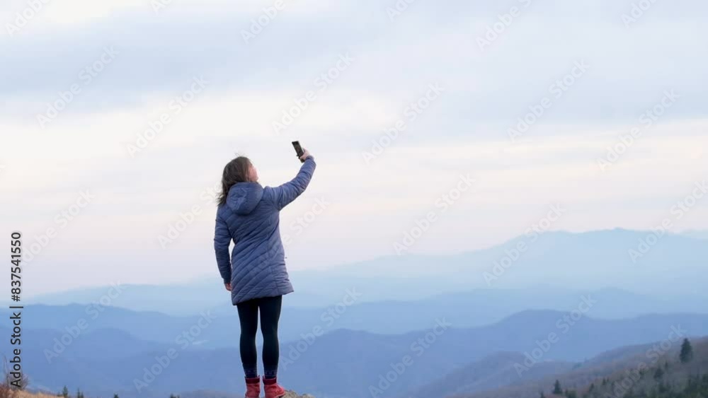A woman stands on a mountain peak, capturing a selfie with her smartphone against the backdrop of an expansive mountain range under a cloudy sky, enjoying the scenic view A woman stands on a mountain peak, capturing a selfie with her smartphone against the backdrop of an expansive mountain range under a cloudy sky, enjoying the scenic view