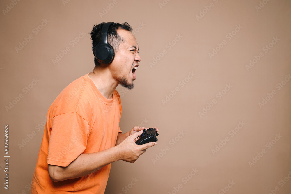 Side view of angry Asian guy screaming while using headphones and joystick losing video games isolated on beige background Side view of angry Asian guy screaming while using headphones and joystick losing video games isolated on beige background