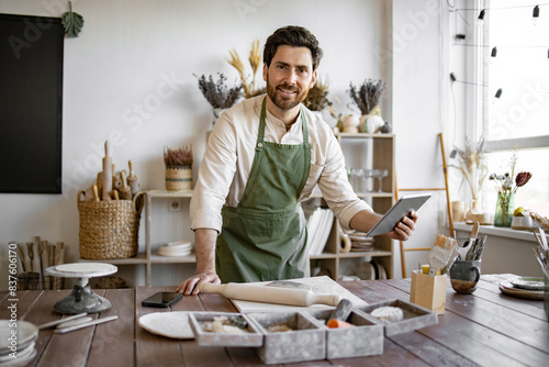 Concept of small business  handmade products and online sales on Internet. Bearded adult man in apron and white shirt viewing an online course on pottery and making ceramic dishes.