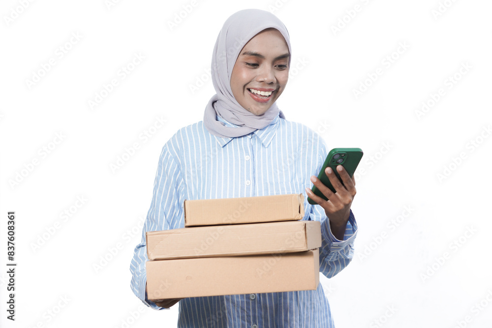 Smiling friendly Asian Muslim Woman in blue white shirt holding package box and use smartphone isolated on white background. Smiling friendly Asian Muslim Woman in blue white shirt holding package box and use smartphone isolated on white background.
