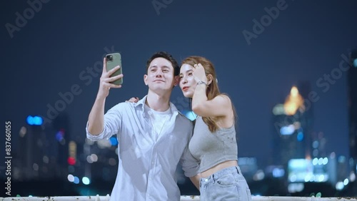 Asian woman and white boyfriend using smartphone taking selfies of night city skyscrapers view on background, travel destination, couples taking selfies at night, 5g network, travel urban city night 