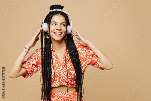 Young smiling happy fun Latin woman she wears orange casual clothes listen to music in headphones look aside on area isolated on plain pastel light beige background studio portrait. Lifestyle concept.