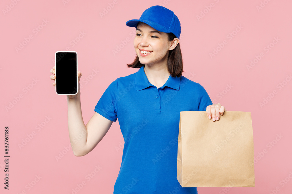 Delivery girl employee woman wears blue cap t-shirt uniform workwear work as dealer courier use mobile cell phone hold brown clear blank paper bag isolated on plain pink background. Service concept. Delivery girl employee woman wears blue cap t-shirt uniform workwear work as dealer courier use mobile cell phone hold brown clear blank paper bag isolated on plain pink background. Service concept.
