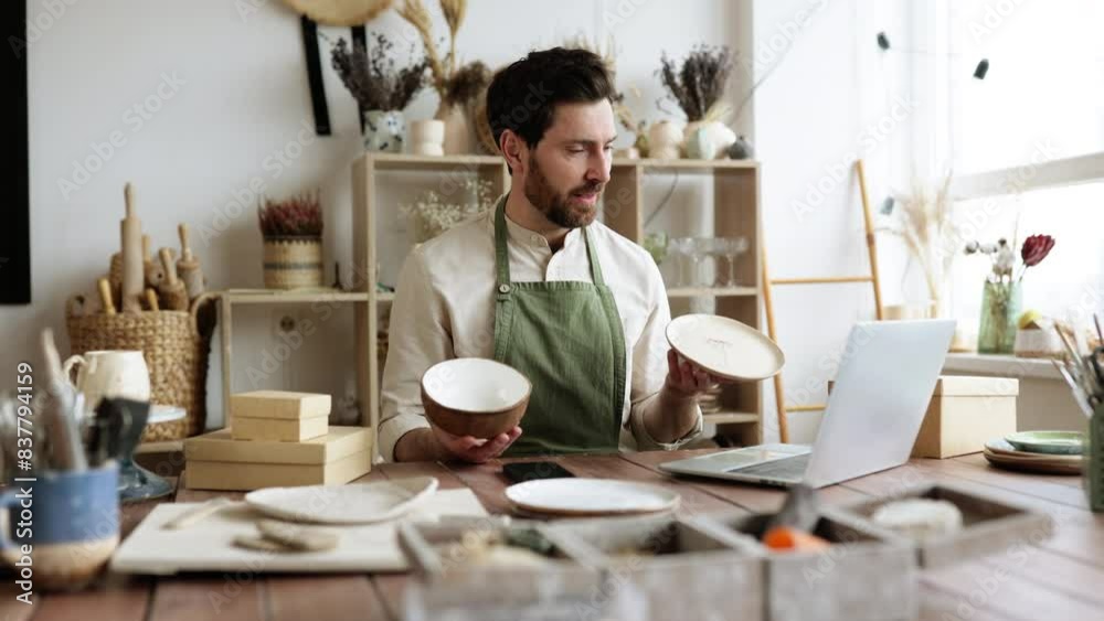 Professional seller checking for no damages on bowl and plate that holding in hands. Attractive bearded man wearing green apron receiving goods at modern decor store. Professional seller checking for no damages on bowl and plate that holding in hands. Attractive bearded man wearing green apron receiving goods at modern decor store.