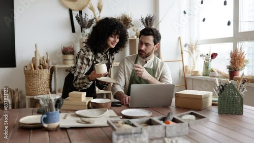 Portrait of mature man and young curly woman creating online catalogue of available goods at decor store. Two workers holding original ceramic products, using laptop during working at shop. photo