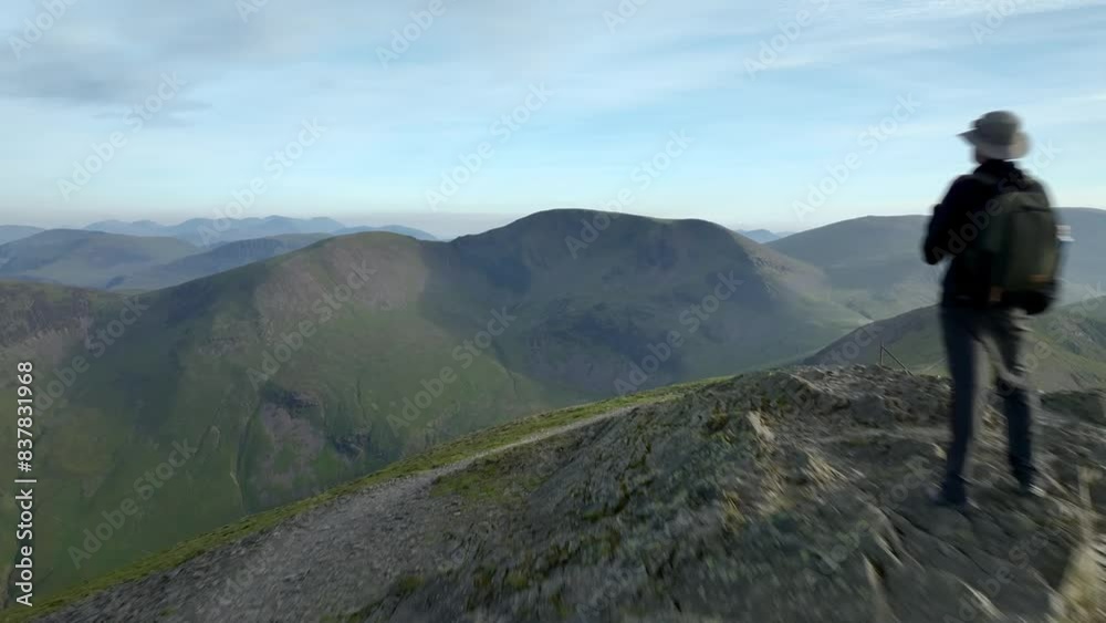 Lone mountain walker on fell summit at golden hour. Camera flypast revealing shadowed deep valley and distant mountains. Grisedale Pike, Lake District, Cumbria, England, UK Lone mountain walker on fell summit at golden hour. Camera flypast revealing shadowed deep valley and distant mountains. Grisedale Pike, Lake District, Cumbria, England, UK
