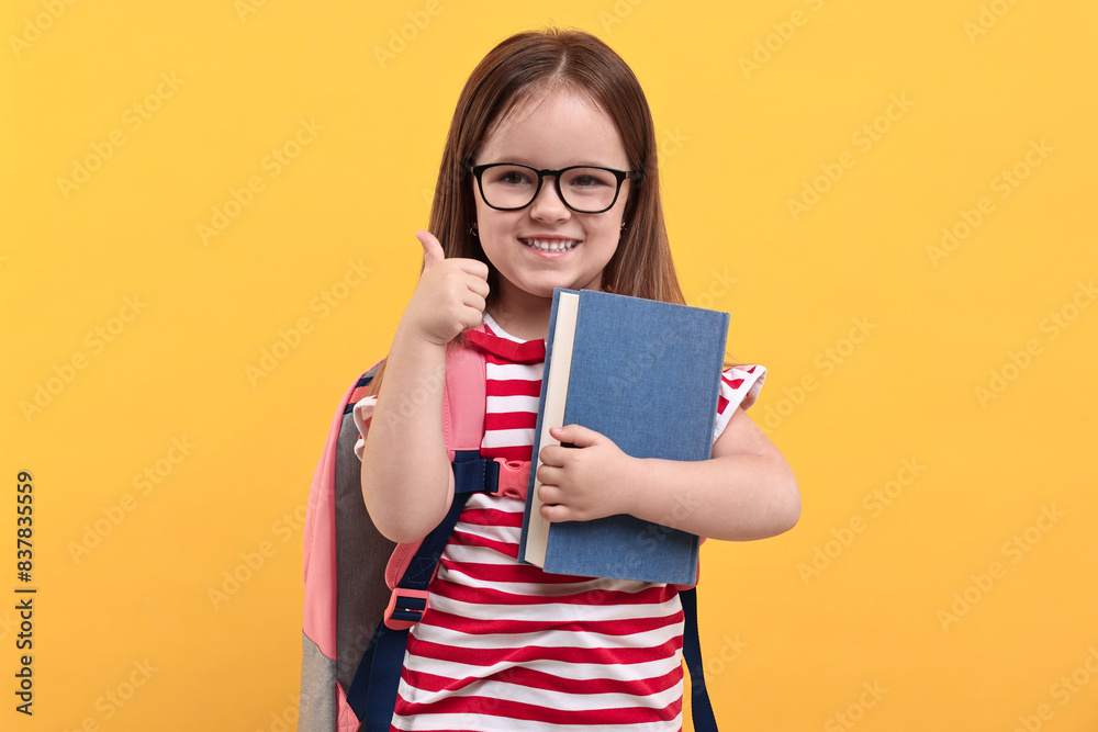 Cute little girl with backpack and book showing thumbs up against orange background Cute little girl with backpack and book showing thumbs up against orange background