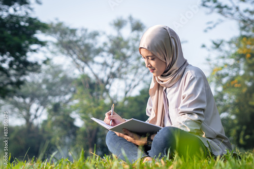 Young Asian muslim woman writing in a notebook while sitting on grass in a sunny morning during summer at the park photo