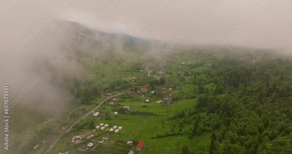 Aerial descending from clouds, revealing a small rural city nestled in a valley between mountains surrounded by green forests
