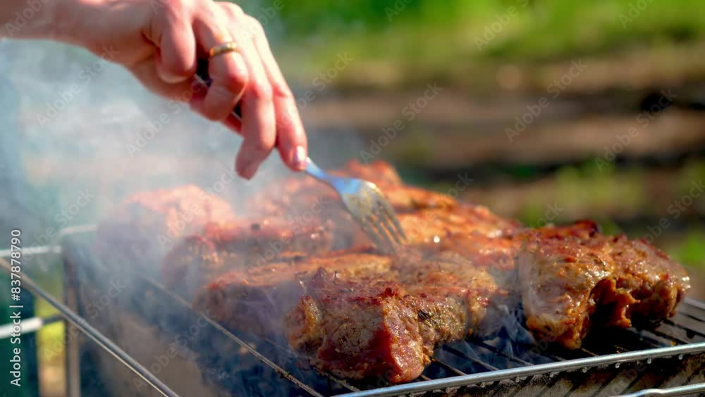 Smoky pieces of meat on grill rack, woman checks doneness of barbecue with fork close-up. Juicy steak with grilling stripes preparing for picnic celebration Smoky pieces of meat on grill rack, woman checks doneness of barbecue with fork close-up. Juicy steak with grilling stripes preparing for picnic celebration