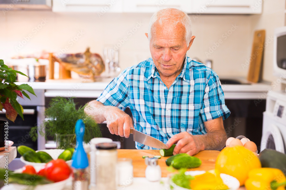 Elderly man cuts vegetables for salad at the table in the kitchen Elderly man cuts vegetables for salad at the table in the kitchen