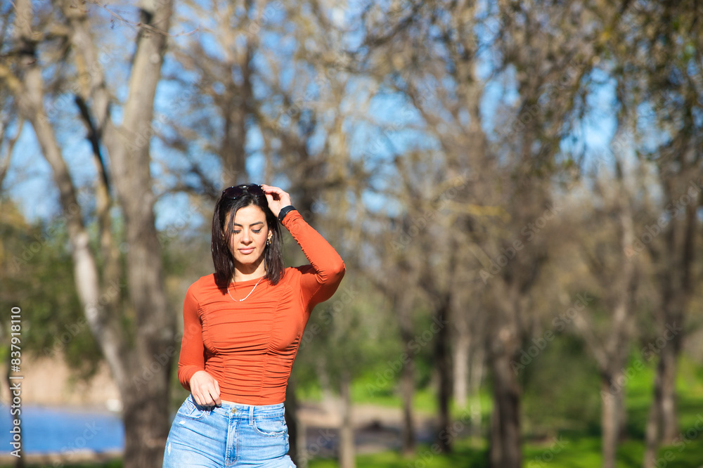 Young and beautiful brunette woman dressed in casual clothes and sunglasses, walks along a path between deciduous trees in autumn, in the background a lake. Young and beautiful brunette woman dressed in casual clothes and sunglasses, walks along a path between deciduous trees in autumn, in the background a lake.