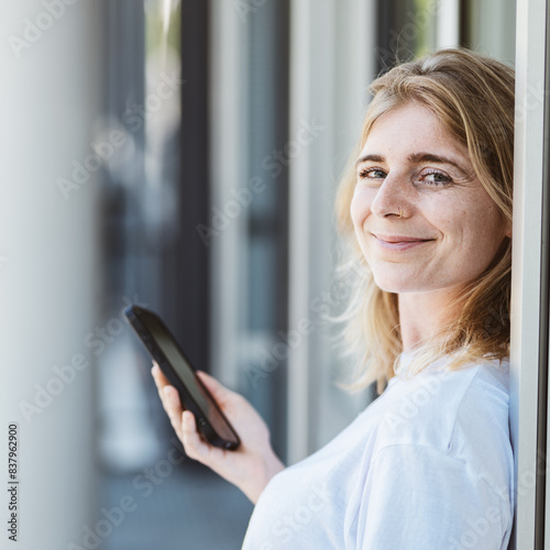 A cheerful woman with a smartphone, seen leaning against a wall and smiling. The photo depicts connectivity and happiness.