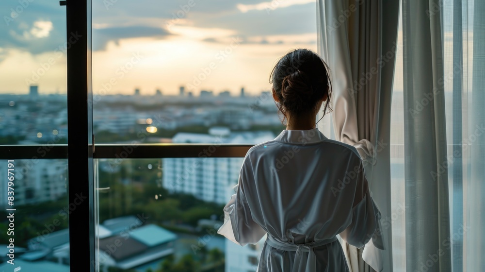 A woman in a white robe stands in front of a window looking out at the city. The scene is peaceful and serene, with the woman taking in the view of the city skyline A woman in a white robe stands in front of a window looking out at the city. The scene is peaceful and serene, with the woman taking in the view of the city skyline