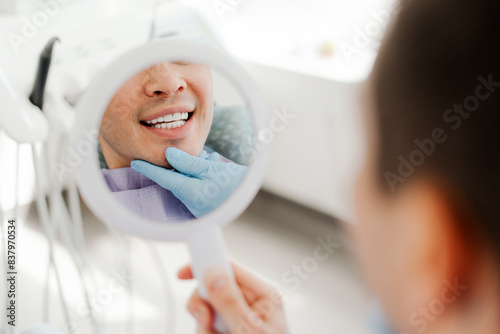Smiling attractive young man  patient holding mirror looking at teeth  treatment in modern dentistry