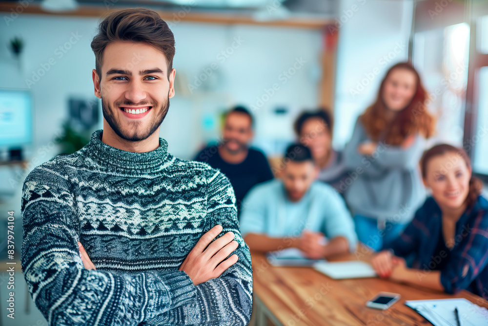 Obraz premium Smiling man in office with colleagues. Smiling young man in a cozy sweater stands confidently in a modern office with his diverse team in the background.