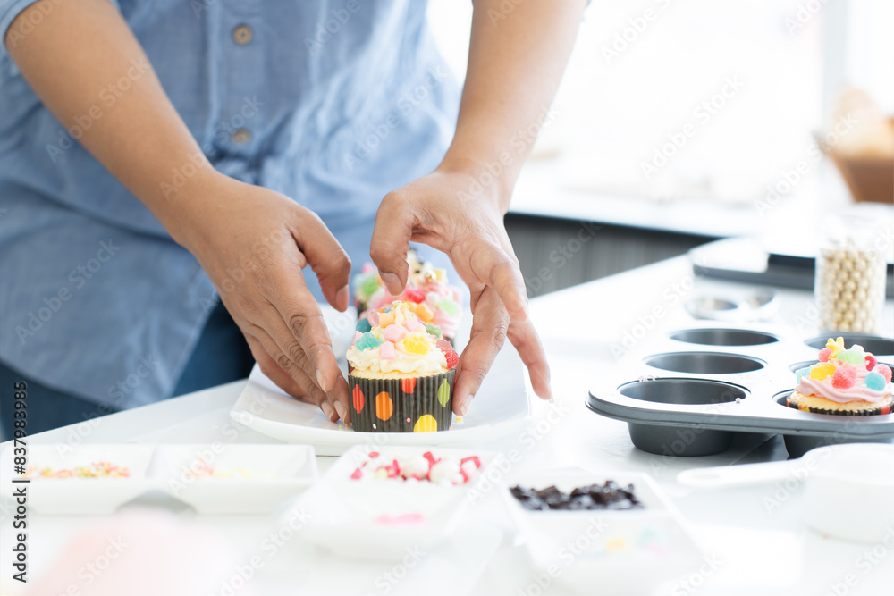 Selective focus on woman hands holding fresh baked cupcakes with decorations from baking tray placing on white plate on table at kitchen at home