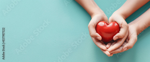 Adult and child hands holding a red heart on an aqua background symbolize heart health, donation, corporate social responsibility, World Heart Day, World Health Day, and Family Day.
