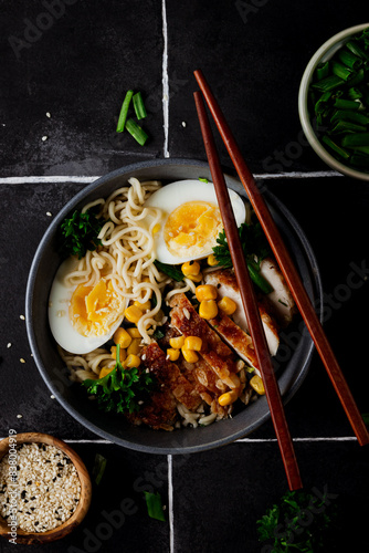 A delicious and hearty bowl of chicken ramen with a boiled egg, fresh green onion, and sweet corn, served over a bed of noodles in a flavorful broth. Black tile background
