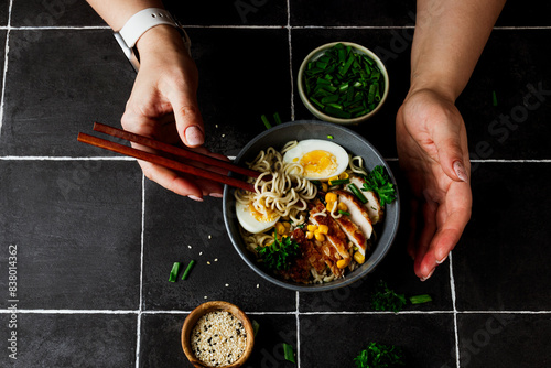 Bowl of chicken ramen with chopsticks and female hands holding them, ready to dig into the delicious broth, noodles, and toppings. The black tile background adds a dramatic touch to the image.