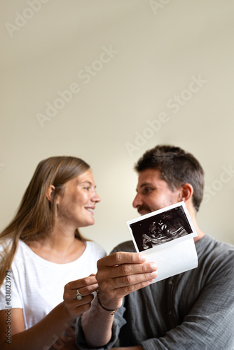 Vertical portrait of pregnant couple looking at each other in love while holding baby ultrasound.Selective focus on hand