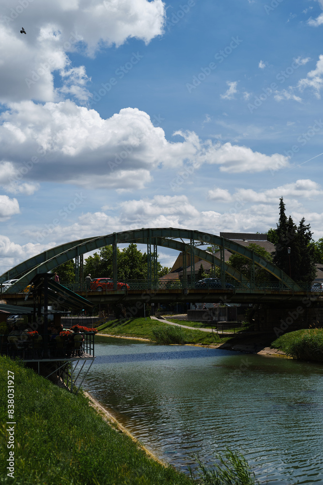 Obraz premium A small iron arch bridge over the river, on which cars and pedestrians drive. The canal is in the city center, people are having lunch at the cafe on the embankment. Town in Serbia is Zrenjanin.