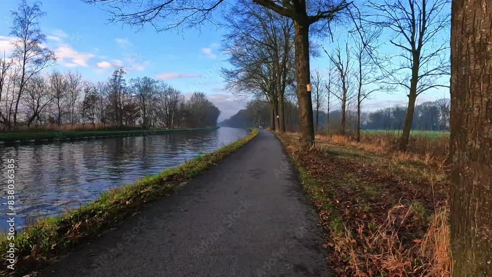 A serene, peaceful setting with a paved pathway along a calm canal, lined with trees and autumn leaves A serene, peaceful setting with a paved pathway along a calm canal, lined with trees and autumn leaves