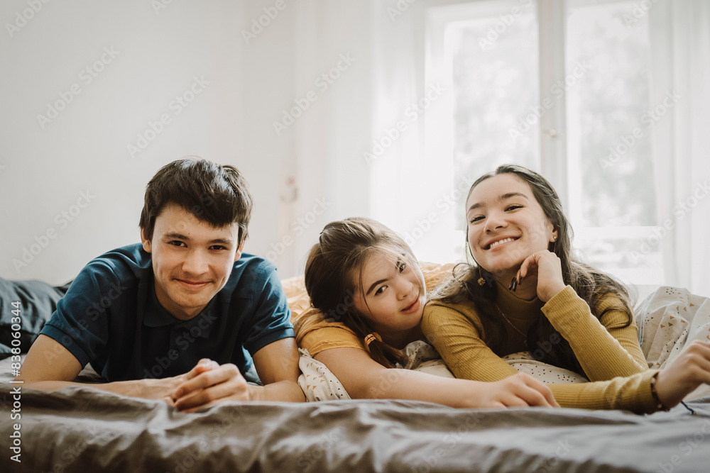 Portrait of smiling woman and teenage boy lying down on bed with down syndrome sibling at home Portrait of smiling woman and teenage boy lying down on bed with down syndrome sibling at home