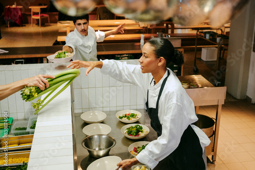 Female chef taking celery while male colleague passing salad bowl in commercial kitchen photo