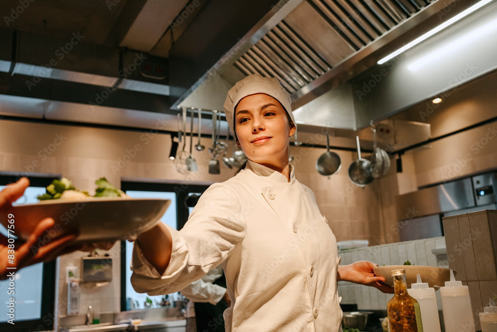Female chef passing salad bowl to colleague working in illuminated kitchen Female chef passing salad bowl to colleague working in illuminated kitchen