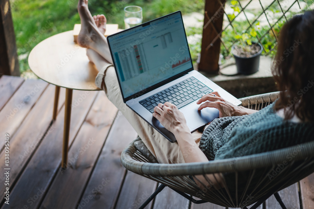Young adult Caucasian woman sitting on cottage terrace using laptop in summertime, online work