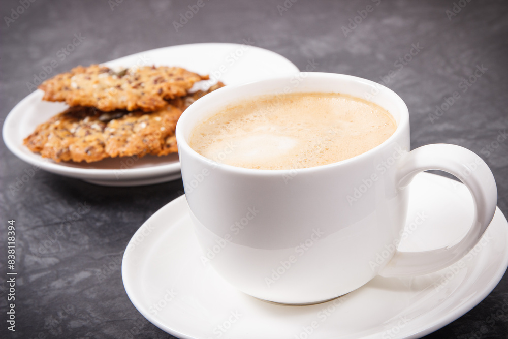 Fototapeta premium Cup of coffee with milk and fresh baked oatmeal cookies with honey and healthy seeds on white plate. Delicious crunchy dessert