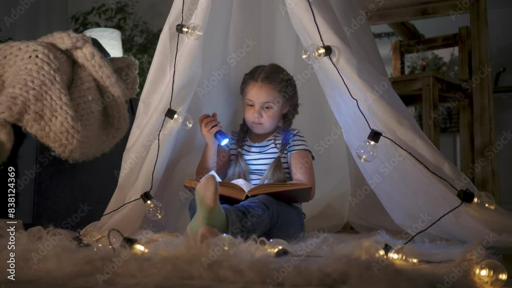 Little girl with lantern reads in tent, child plays at home in imaginative setting. Cozy den, child plays at home, engrossed in book in tent. Child plays at home, nestled in tent with book and lantern