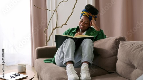 African American girl sits on couch alone, holding notebook and writing thoughts down in diary. Female black student taking notes or young writer working on a new story. She ponders and thinks over.