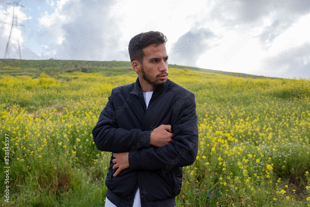 A man in a black jacket and dishdasha stands in a vibrant field of yellow flowers and green hills, arms crossed for warmth, under a cloudy spring sky, feeling cold A man in a black jacket and dishdasha stands in a vibrant field of yellow flowers and green hills, arms crossed for warmth, under a cloudy spring sky, feeling cold