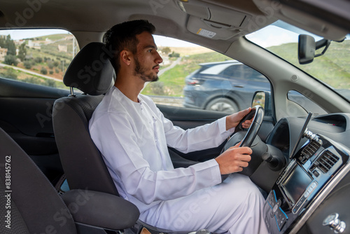 A cheerful young man with a beard, wearing a traditional Middle-Eastern outfit, smiles confidently while seated in his car. He appears to be enjoying a drive through a scenic countryside © nooh