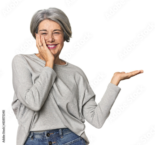Caucasian mid-age female on studio background holds copy space on a palm, keep hand over cheek. Amazed and delighted.