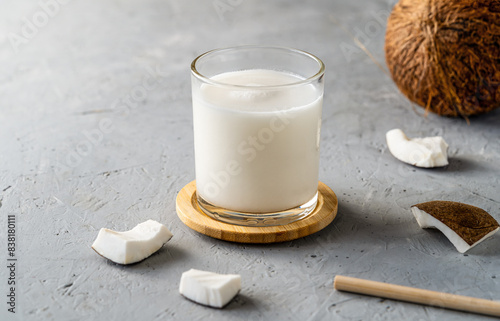 A glass of fresh coconut milk on a wooden coaster, surrounded by coconut chunks and a whole coconut on a textured gray surface.