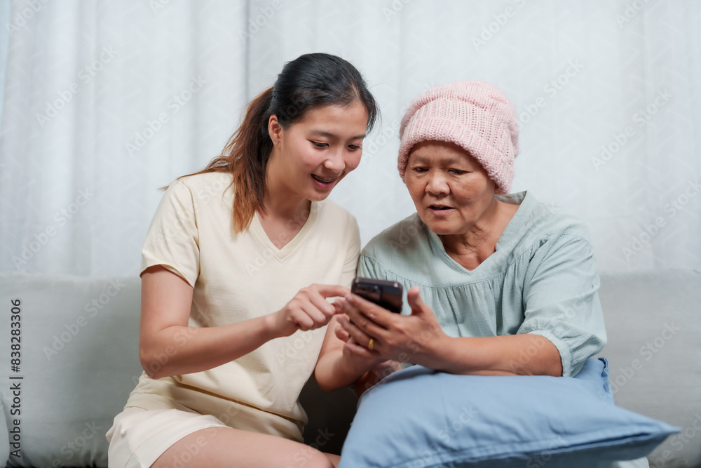 Delight visible on faces of elderly and young women as they view a smartphone, seated comfortably at home. Mirth shared between aged female in azure top and young counterpart over phone visuals Delight visible on faces of elderly and young women as they view a smartphone, seated comfortably at home. Mirth shared between aged female in azure top and young counterpart over phone visuals