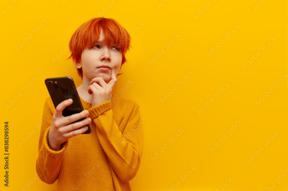 pensive red-haired child boy using smartphone and planning on yellow isolated background, puzzled teenager winner with orange hairstyle thinks and imagines and looks away pensive red-haired child boy using smartphone and planning on yellow isolated background, puzzled teenager winner with orange hairstyle thinks and imagines and looks away