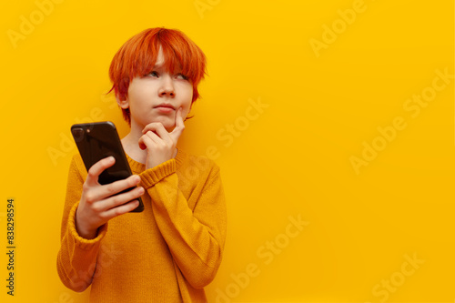 Fotografie pensive red-haired child boy using smartphone and planning on yellow isolated ba
