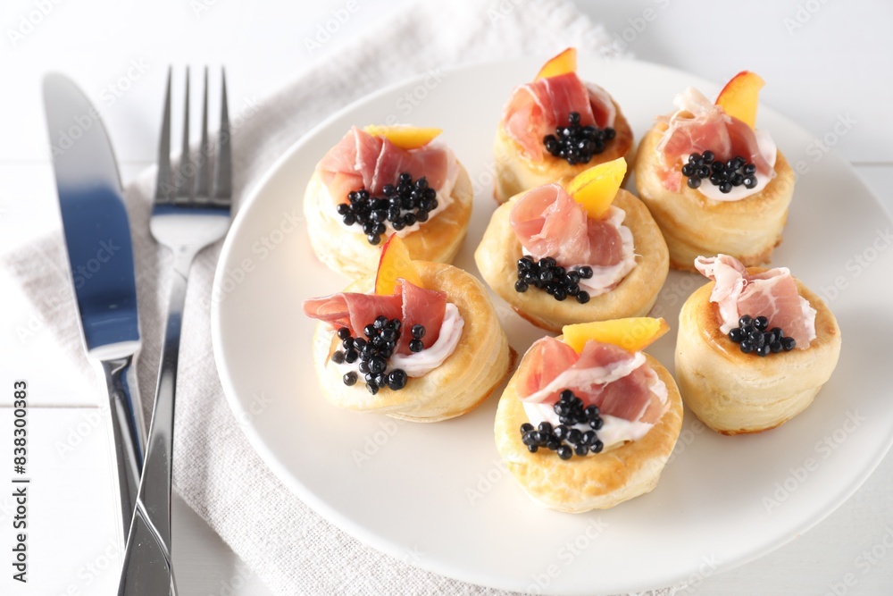 Delicious puff pastry with cream cheese, ham, black caviar, peach and cutlery on white wooden table