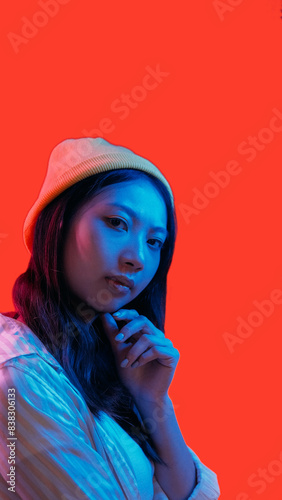 Neon people. Hipster portrait. Blue pink color light pensive skeptic girl in beanie posing touching chin isolated on red empty space background.